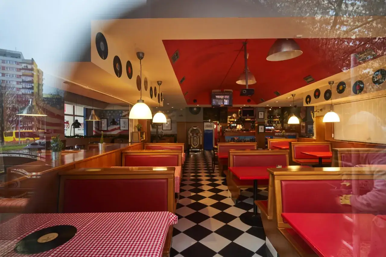 Wnętrze w stylu retro z czerwonymi kanapami i czarno-białą podłogą w szachownicę. W tle widać jukebox i dekoracje z płyt winylowych, idealne dla Queens Street Bar.