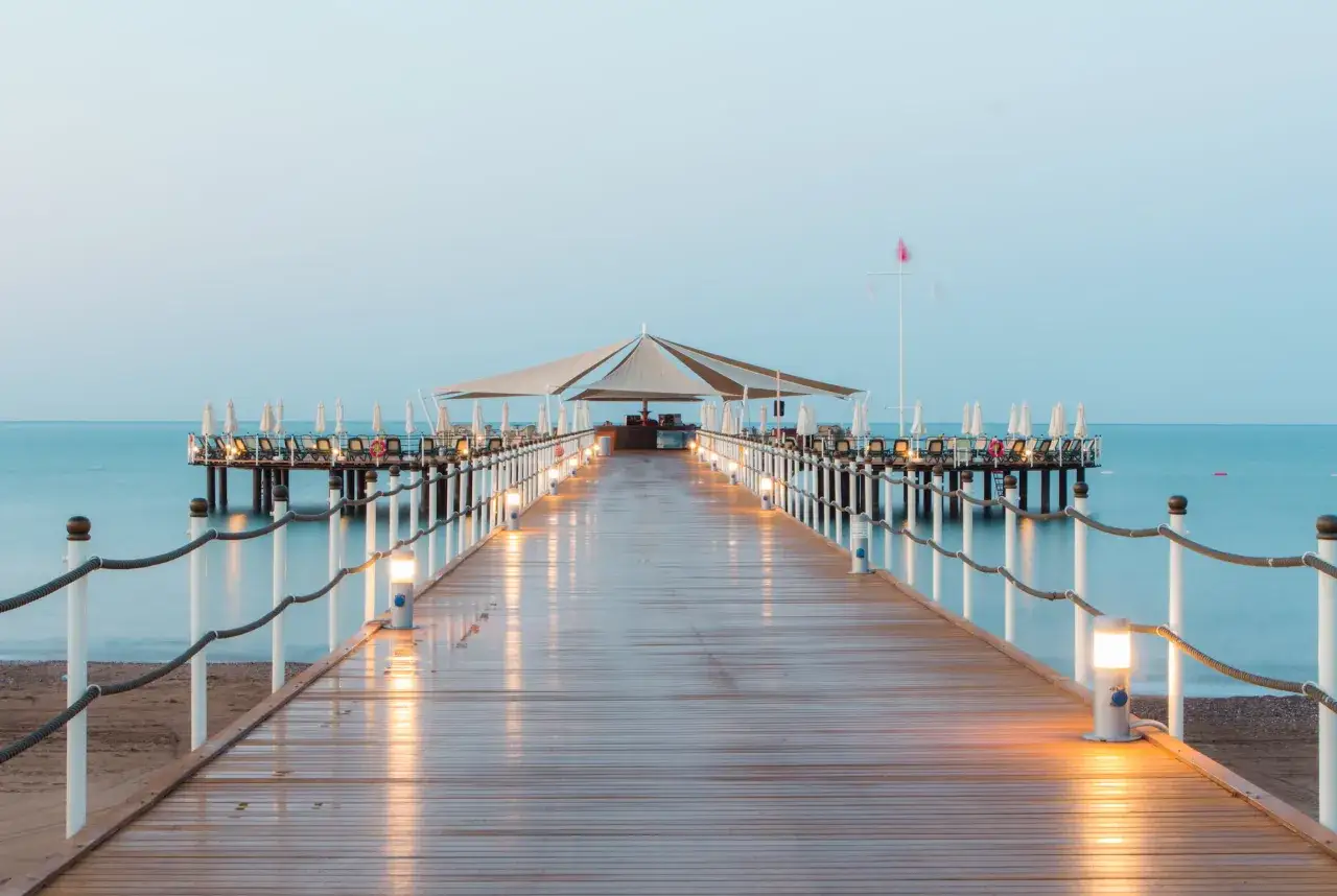Drewniane molo z oświetleniem prowadzi do pomostu z leżakami i parasolami. Idealne miejsce na relaks w Orange County Belek.